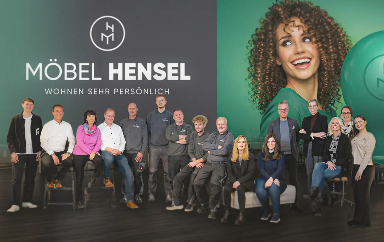 Gruppe von Personen steht und sitzt vor Wand mit Schriftzug Möbel Hensel und Wohnen sehr persönlich sowie großem Portrait rechts